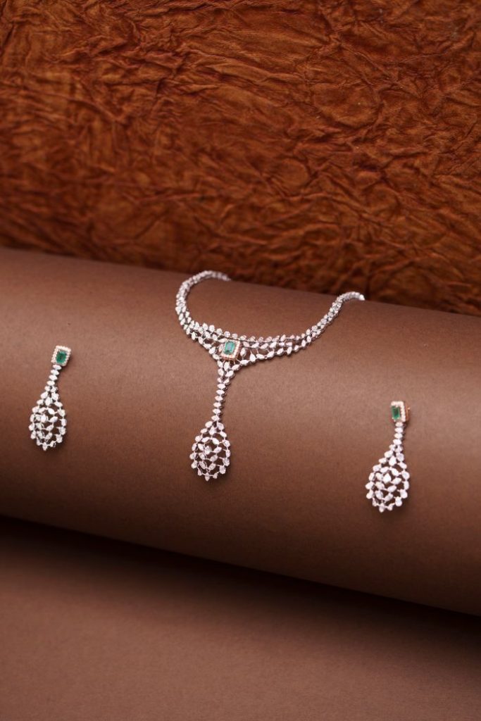Exquisite diamond necklace and earrings with emerald accents displayed on a rich brown background.
